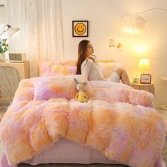 a woman sitting on a bed covered in pink and orange fluffy blankets with a stuffed animal next to her