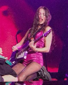 a woman in a pink dress playing an electric guitar on stage with a man sitting next to her