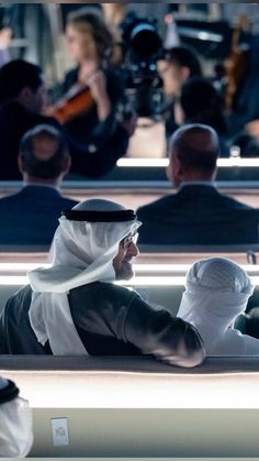 two men in white headscarves are sitting on a bench and one man is playing the violin