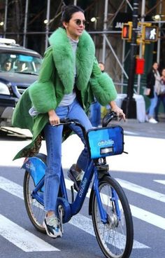 a woman is riding her bike down the street wearing a green coat and blue jeans