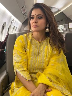 a woman sitting on an airplane seat wearing a bright yellow outfit with gold jewelry and earrings