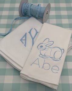 two white towels with blue monogrammed letters and a bunny on them are sitting on a checkered tablecloth