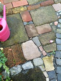 a pink watering can sitting on top of a cobblestone road next to flowers