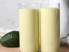 two glasses filled with yellow liquid next to an avocado