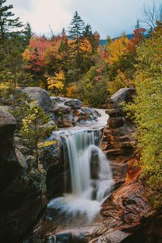 a waterfall in the middle of a forest surrounded by rocks and colorful trees with fall foliage