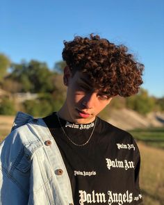 a young man with curly hair wearing a black shirt and denim jacket standing in a field