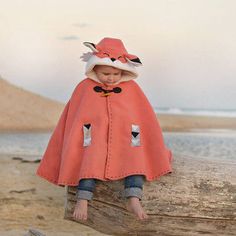 a little boy sitting on top of a wooden log wearing a red cape and hat