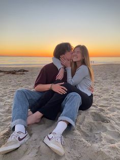 a man and woman sitting on the beach kissing each other while the sun sets in the background