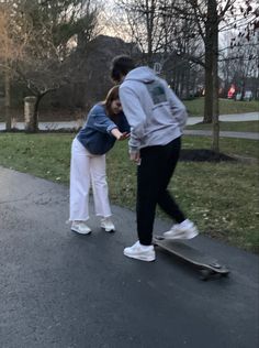 two people standing on a skateboard in the middle of a sidewalk with grass and trees behind them