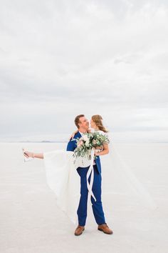 a bride and groom holding each other on the beach in front of an overcast sky