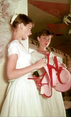 two women in white dresses are holding red ribbons and hats while one woman is looking at the mirror