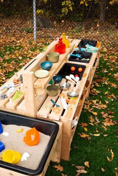 an outdoor play table with bowls and cups on it in the grass next to fallen leaves