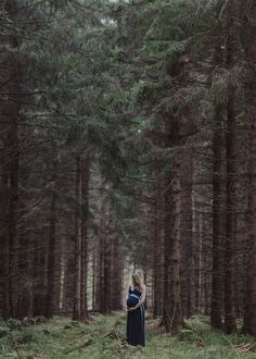 a woman standing in the middle of a forest with tall pine trees behind her and looking at the camera