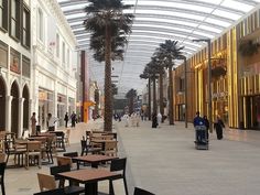 people are walking through an indoor mall with tables and chairs on the ground, palm trees in the background