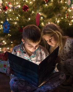 two children sitting in front of a christmas tree looking at an open book with lights on it