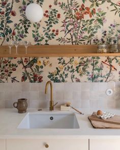 a white sink sitting under a wooden shelf next to a wall covered in floral wallpaper