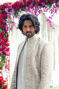 a man with long hair wearing a white outfit and standing in front of red flowers