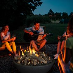 three people sitting around a fire pit playing guitar and drinking wine at night with the lights on