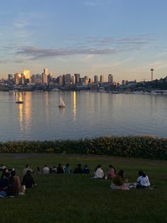 people are sitting on the grass by the water watching boats sail in the bay at sunset