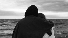 two people are sitting on the beach looking out at the ocean and one person is wearing a hooded jacket