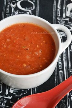 a white bowl filled with tomato soup next to a red spoon on a black and white tablecloth