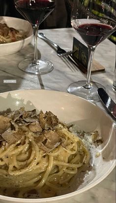 a bowl of pasta with mushrooms and wine glasses on the table in front of it