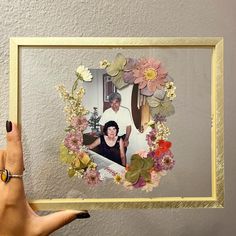 a woman holding up a framed photo with flowers and leaves in the frame on her left hand