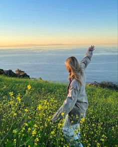 a woman is standing in a field with her arms up and looking at the ocean