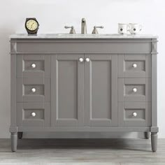 a gray bathroom vanity with two sinks and a clock on the wall above it in a white room