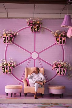 a man sitting in a chair next to a wall with flowers on it and hanging planters