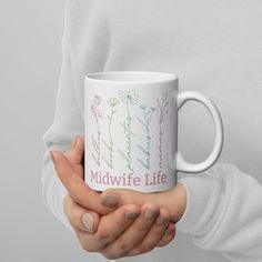 a woman holding a white coffee mug with flowers on the side and midwife life written on it