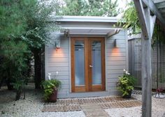 a small gray house with wooden doors and plants in the front yard, surrounded by trees