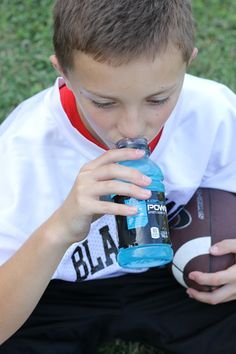 a young boy sitting on the grass drinking from a water bottle while holding a football