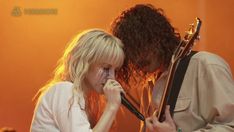 a man and woman are playing guitar together in front of an orange wall with their faces painted white