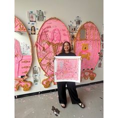 a woman standing in front of a wall with pink artwork on it and holding up a framed print