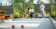 two women are playing bowls in an outdoor bowling alley with wooden benches and picnic tables