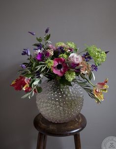 a vase filled with lots of flowers sitting on top of a wooden stool next to a wall