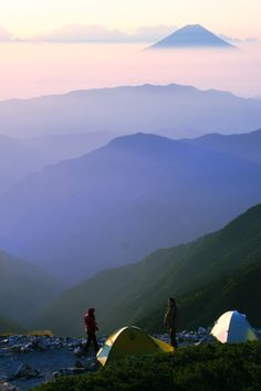 two people standing on top of a mountain with tents in the foreground and mountains in the background