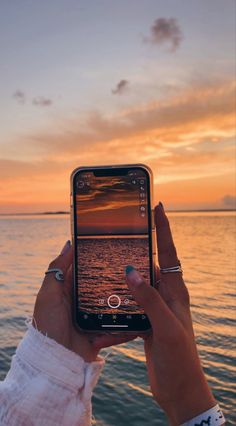 a woman holding up her cell phone in front of the ocean at sunset or sunrise