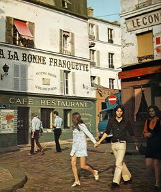 two people are holding hands while walking down the street in front of some shops and buildings