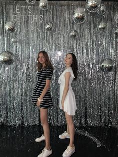 two women standing next to each other in front of a wall with shiny balls on it