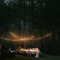 a group of people sitting around a table in the woods at night with lights strung over them