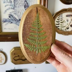 a hand holding a wooden brooch with a green leaf on it in front of framed pictures