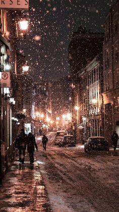 two people walking down a snowy street at night in the city with snow falling on them