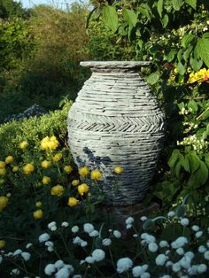 a large gray vase sitting in the middle of a garden filled with yellow and white flowers