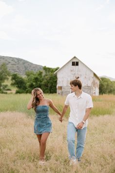 a man and woman holding hands walking through tall grass in front of an old barn