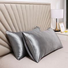 two silver pillows sitting on top of a bed next to a nightstand with a lamp