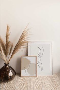 two framed pictures sitting on top of a table next to a vase with dry grass in it