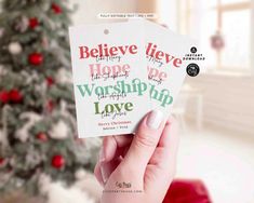 someone holding up two christmas cards in front of a christmas tree with the words believe, hope, and worship