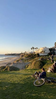 people are sitting on the grass by the beach with their bikes in front of them